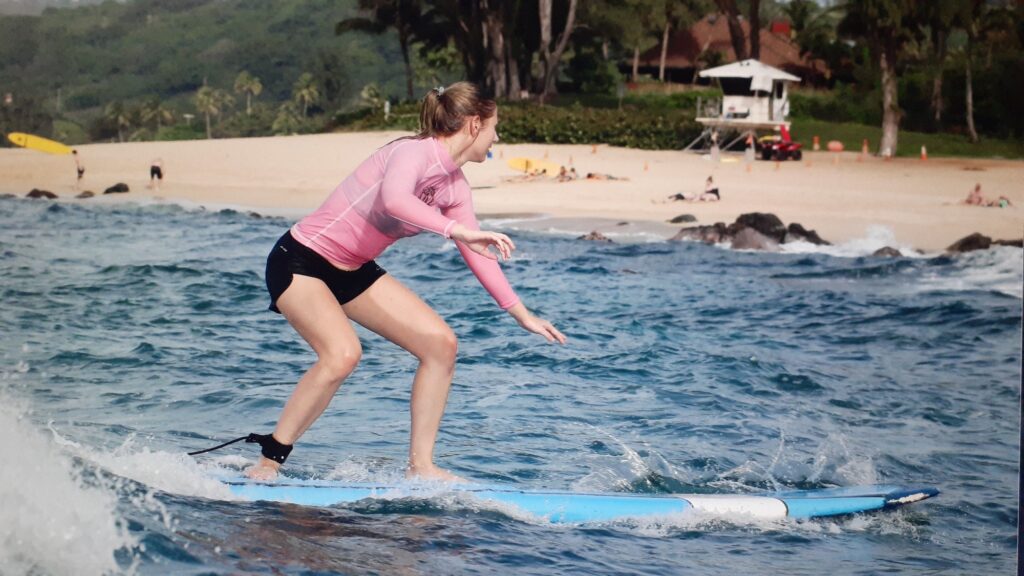 Beginner surfer in a pink long-sleeve rash guard and black shorts crouching on a blue soft-top surfboard, riding a small wave with an ankle leash on, with a sandy beach, rocks, and trees behind.